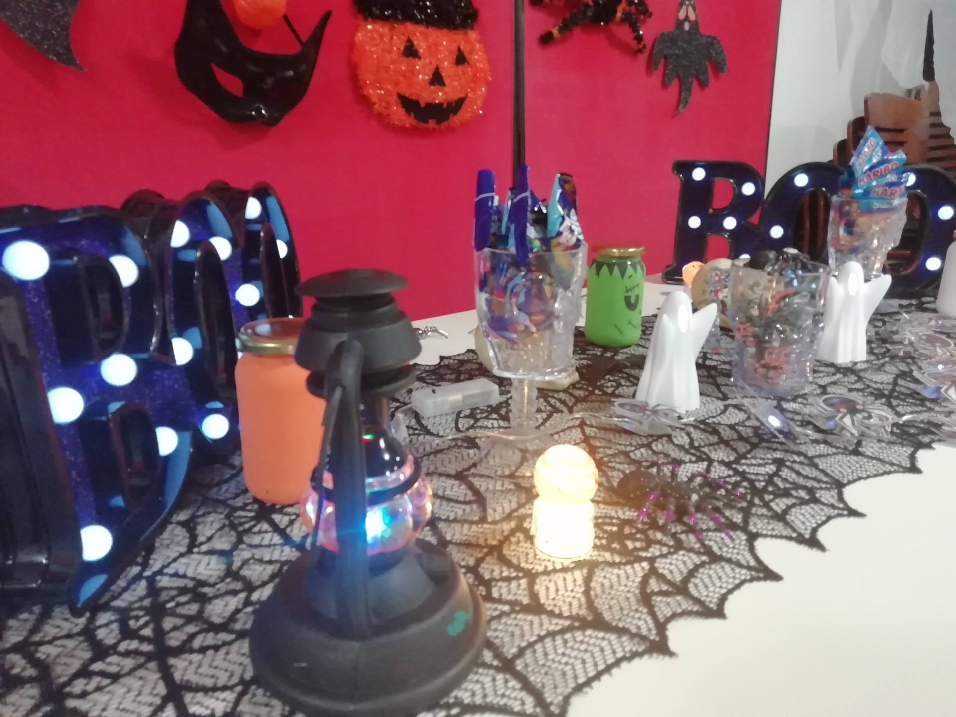 A selection of Halloween decorations on a Halloween tablecloth on a table. There is some lights in the shape of a pumpkin or the word 'Boo!'