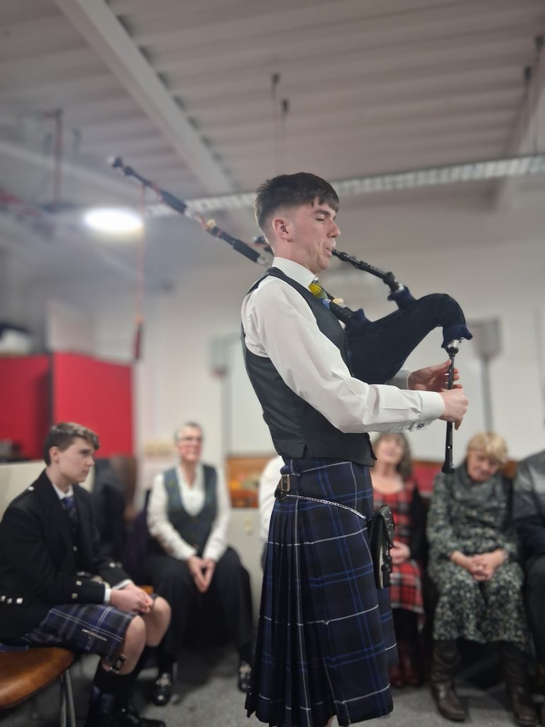 A bagpiper stood playing in The Devil's Porridge Museum's education room. Some people sit around the room listening.