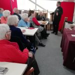 Members of The Devil's Porridge Museum's Cordite Club sat around three tables looking at Tom, who is stood talking to them.