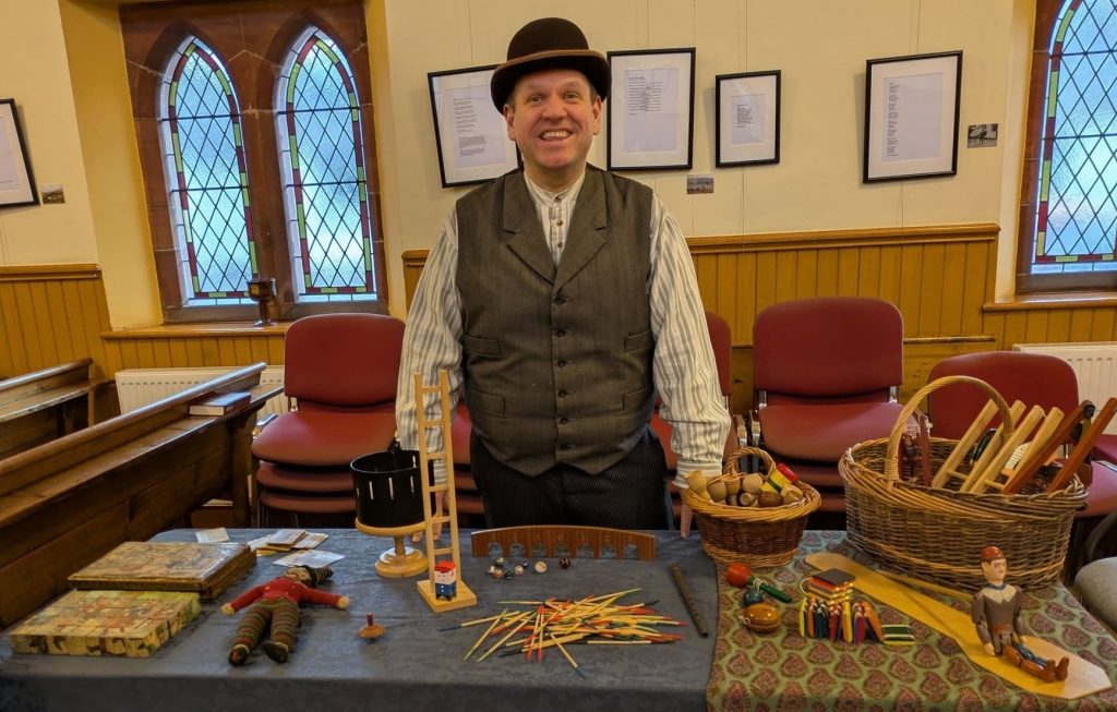 A table full of traditional toys, such as Jacob's Ladder and pick up sticks. The Devil's Porridge Museum's Youth and Community Engagement Officer, Tom, is stood behind the table. He looks happy.