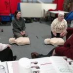 Two CPR models on the floor with two members of The Devil's Porridge Museum's team beside them. Two people are sat at at table in front of this.