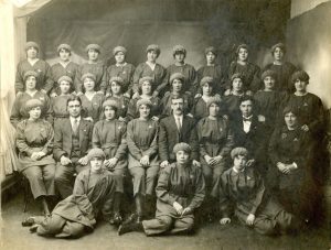 Munitions workers in uniform stood and sat in four rows. This is a black and white photograph from the past.