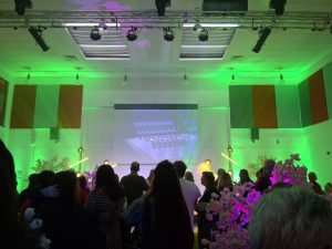 A audience stand in front of a projected screen in a room with green lighting.