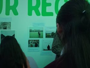 Two people stood looking at some photos of people in different local places on a wall.