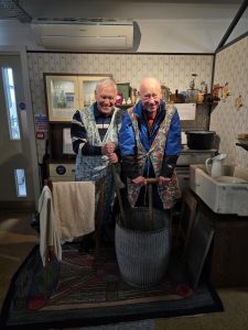 Two men dressing up in 1940s style aprons and posing as though they are washing clothing in The Devil's Porridge Museum's 1940s house display.