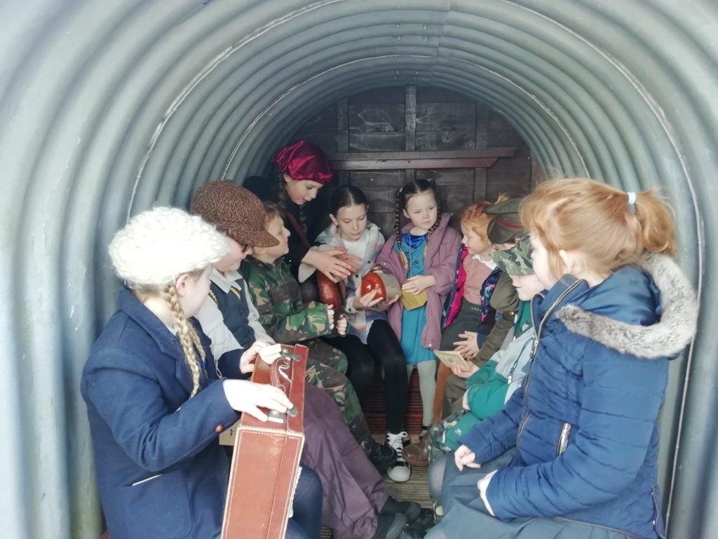 Some children gathered in an Anderson Shelter. Some are holding suitcases.