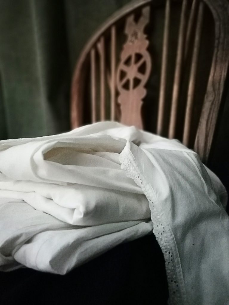 Some cotton blouses folded up on a wooden chair. This chair dates from World War One. There is a dark green curtain in the background.