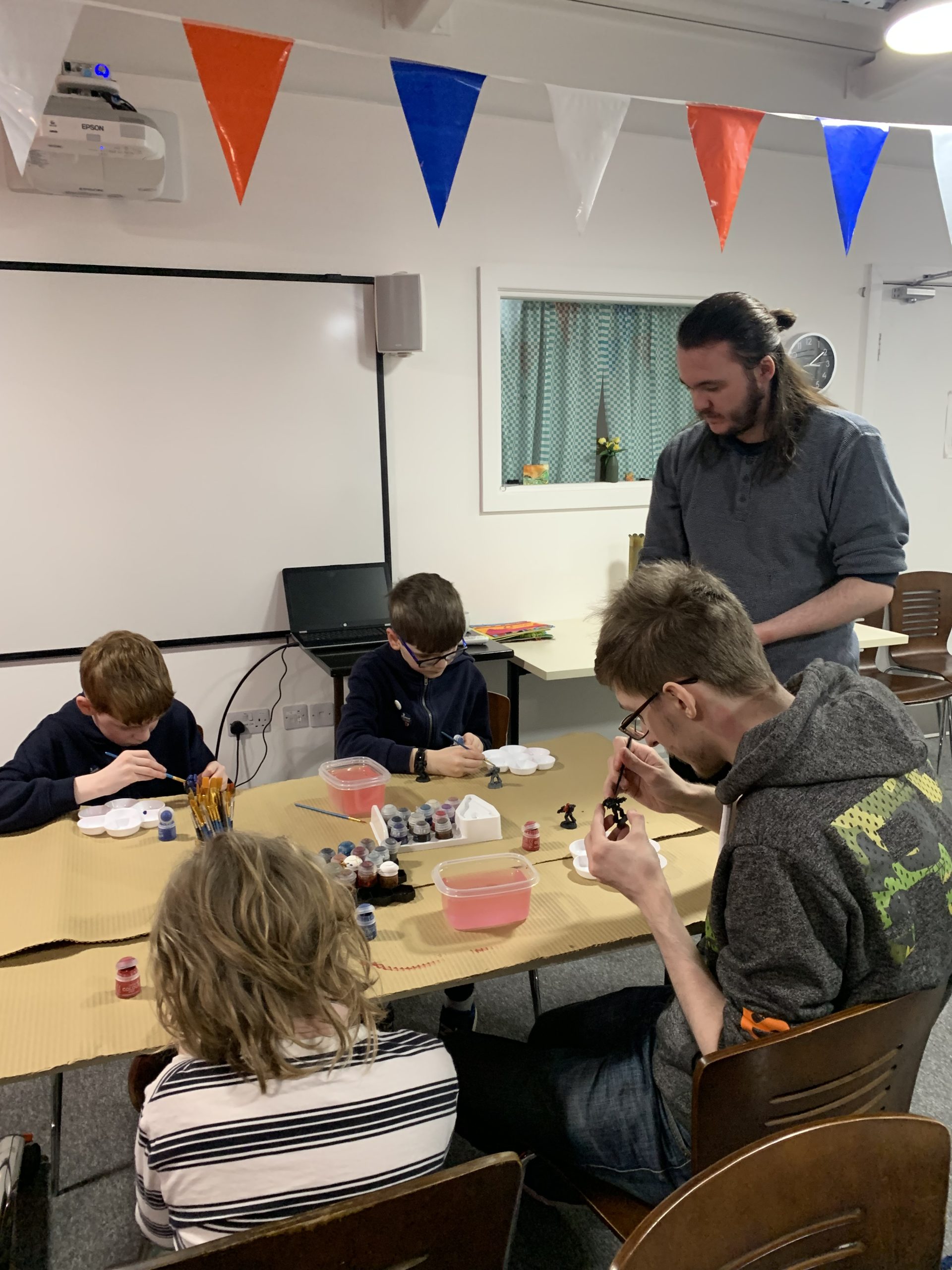 Some young people sat around a table painting Warhammer miniatures. A volunteer is stood up at the end of the table and another is sat painting.