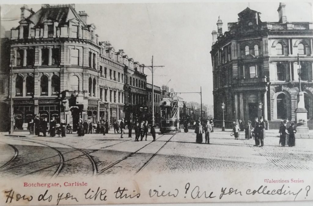 A postcard of Botchergate in Carlisle. There are some tall buildings with people stood on the road between them and some tram lines. This is a black and white photo. Underneath this is a handwritten note which reads "how do you like this view? One you collecting?"