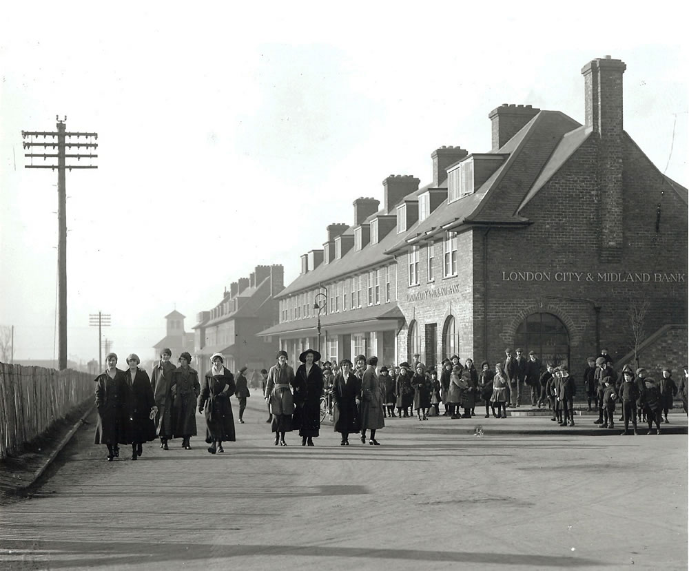 A selection of shops in Gretna with the bank no the near end. Crowds of people are walking down the road and street. This is a black and white photo from the past.