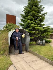 A man dressed as an Air Raid warden holding a shovel and an envelope stood in front of an Anderson shelter outside The Devil's Porridge Museum.