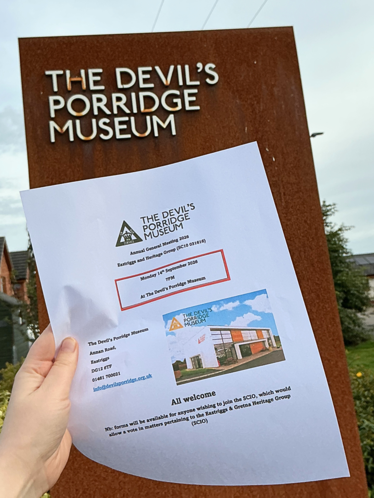 A poster for The Devil's Porridge Museum's Annual General Meeting 2026. This is happening on Monday 14th September 2026 at 7pm at The Devil's Porridge Museum. All welcome. Forms will be available for anyone wishing to join the SCIO, which would allow a vote in matters pertaining to the Eastriggs and Gretna Heritage Group (SCIO). This poster is held up in someone's hand in front of a sign which reads "The Devil's Porridge Museum."
