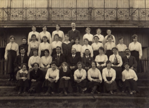 A group of people in arranged in four rows for a photo. Kitty is in the third row from the front, first person on the left. The lady dressed in black beside her is her mother, Rose Ann Palmer. This is a black and white photo from the past.